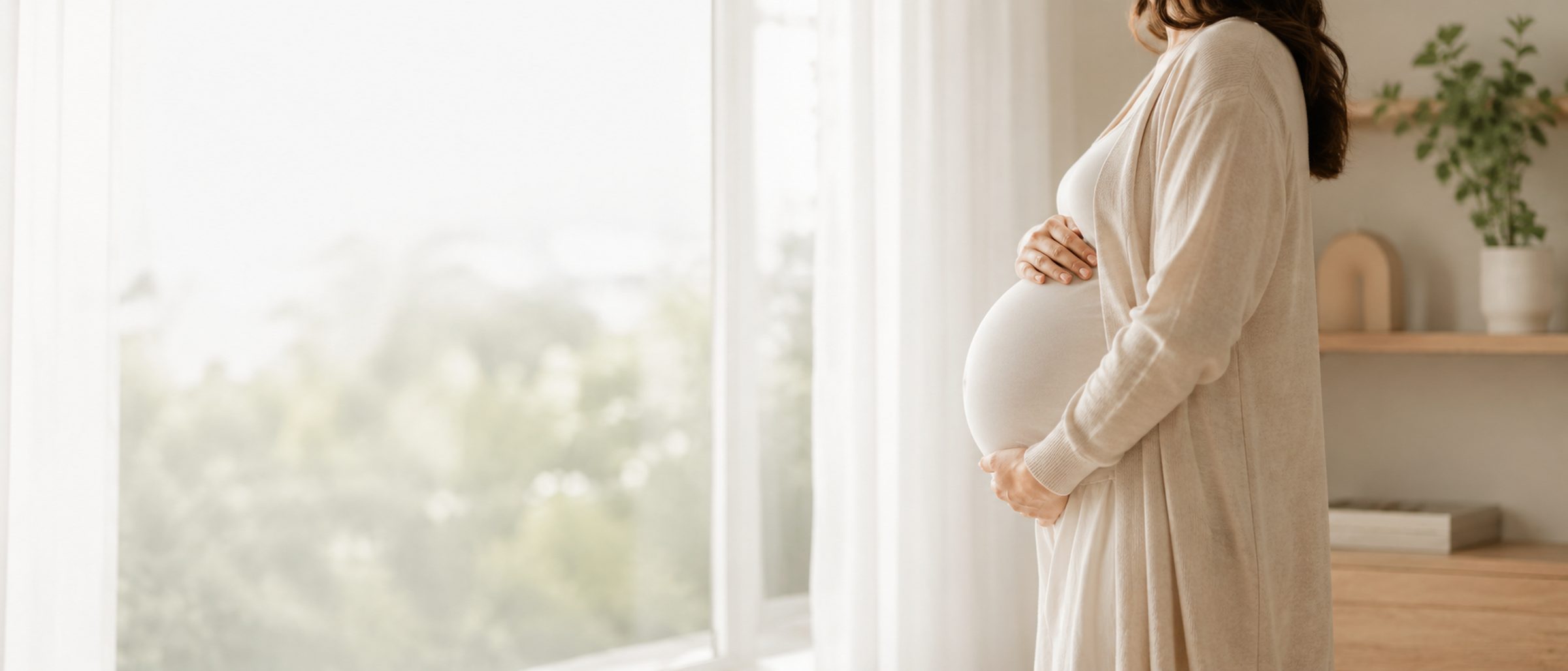 Gestante de perfil junto à janela, com as mãos sobre a barriga